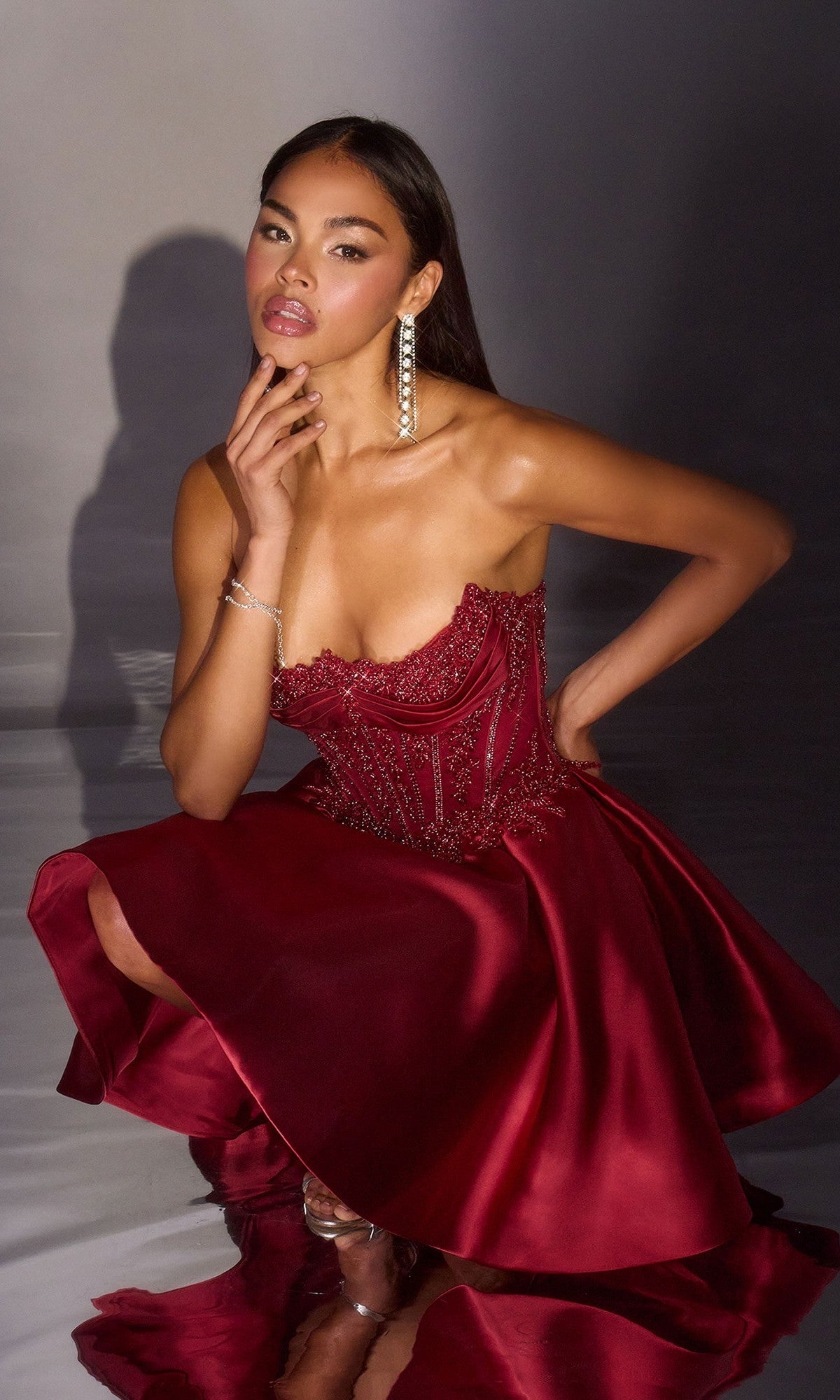 Model in the Ladivine CC502 Short Homecoming Dress featuring a strapless design, deep red satin A-line skirt, and intricate detailing. She poses seated on one knee, looking at the camera with one hand near her chin.